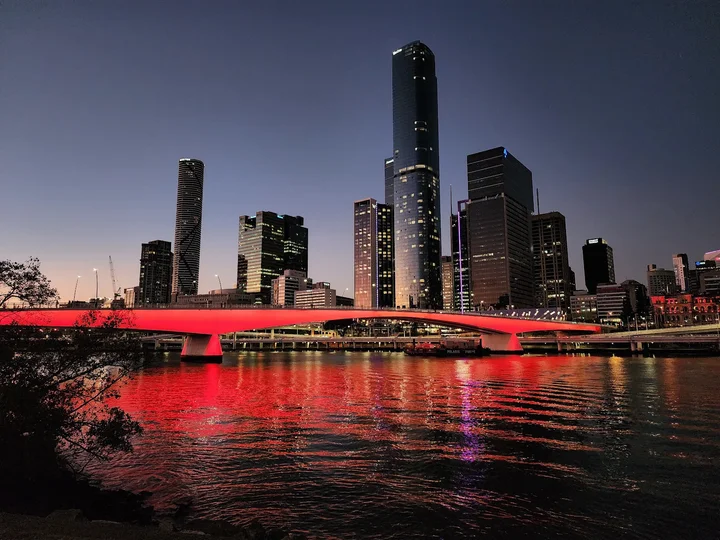 Cityscape at dusk featuring a modern skyline with tall buildings and a brightly illuminated bridge casting red reflections on the river below. The sky is clear, transitioning from light to dark.