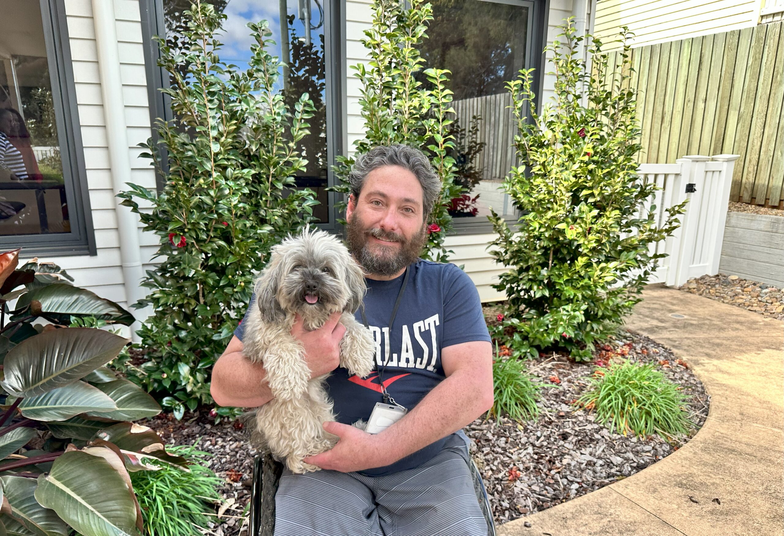 A man with a beard sits in a garden holding a small, shaggy gray and white dog on his lap, smiling at the camera—showing the joy you can bring when you leave a gift in your will. Green bushes and a house with white siding are in the background.