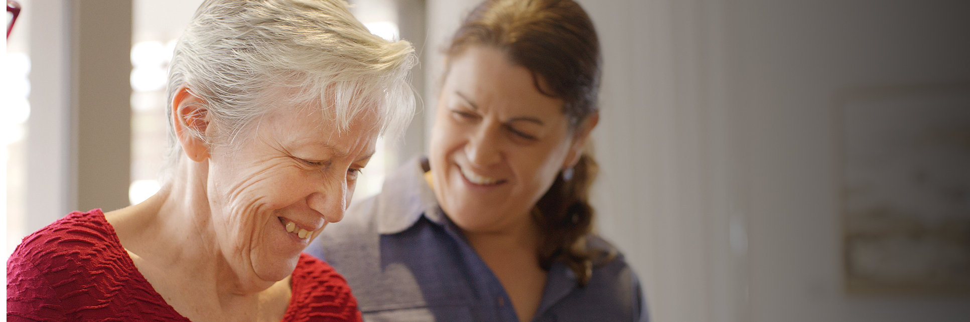 Two women smile warmly at each other indoors. The older woman, with short gray hair and a red top, looks down while the younger woman, with brown hair and a blue shirt, stands behind her, smiling supportively.