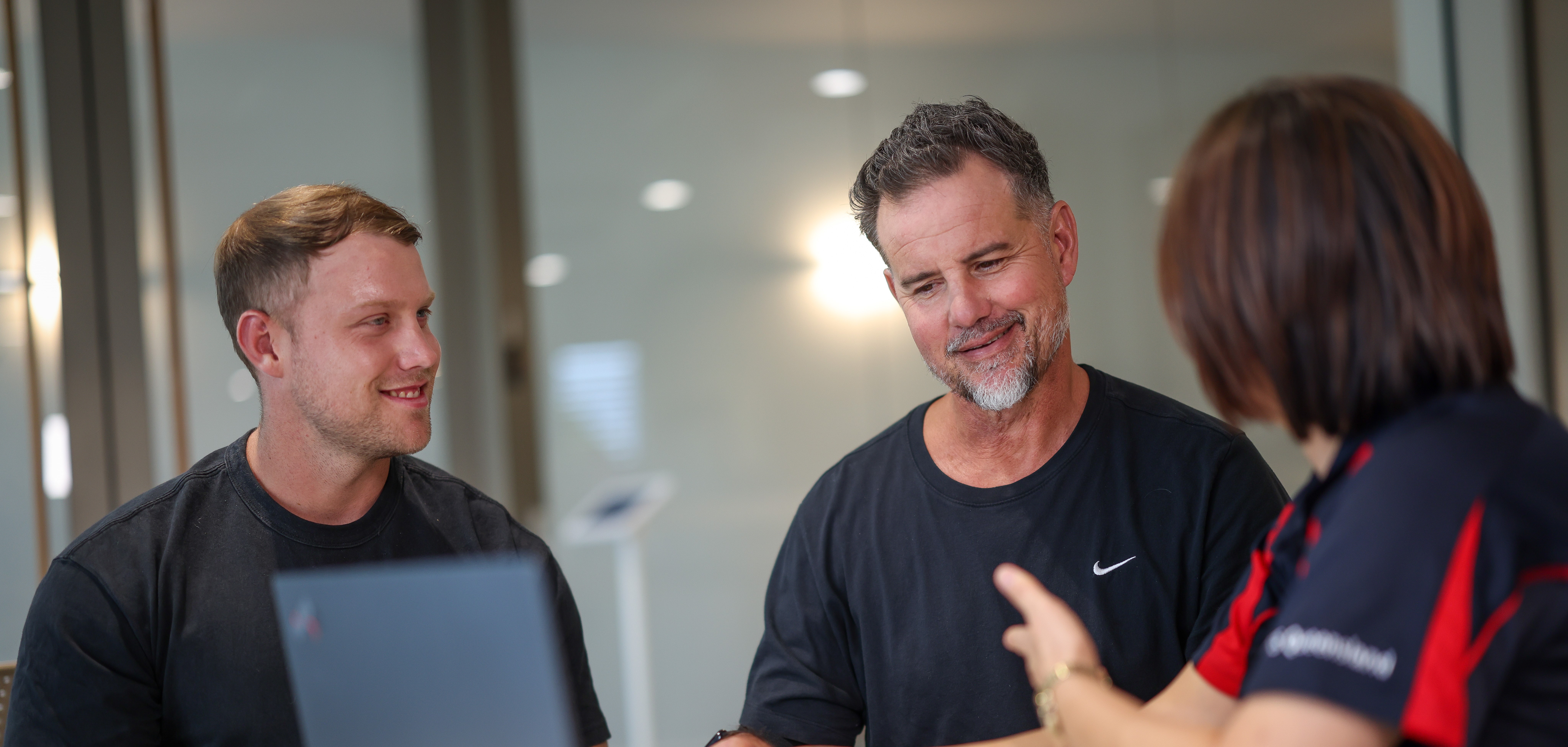 Three people sit at a table having a discussion. Two men face the camera and smile, while the woman in the foreground gestures with her hand. A laptop is open on the table.