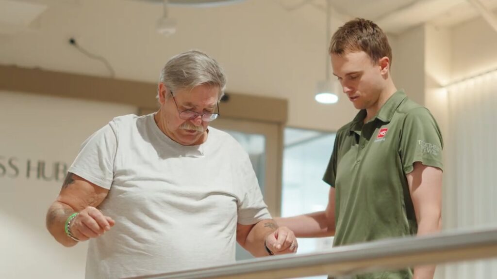 An older man in a white t-shirt receives assistance from a younger man in a green shirt inside a bright indoor setting, possibly a healthcare or rehabilitation facility.