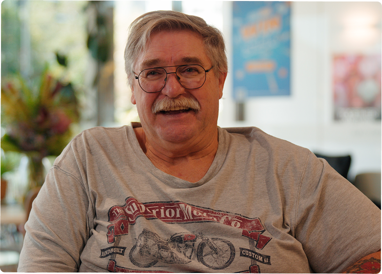 Older man with gray hair, glasses, and a mustache smiles while sitting indoors. He wears a light gray T-shirt featuring a vintage motorcycle design. The background is softly blurred with some plants and posters visible.
