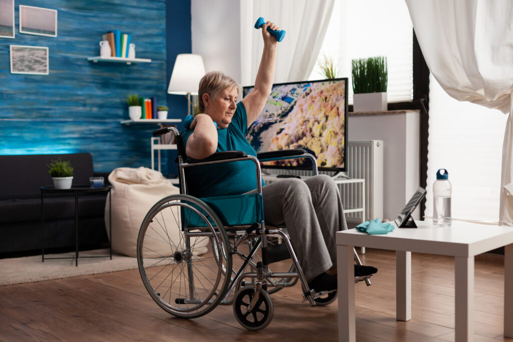 An older adult in a wheelchair lifts a dumbbell at home, exercising in front of a tablet as part of their neurological physiotherapy routine. The room is bright and cozy, with blue walls, plants, and a TV in the background.