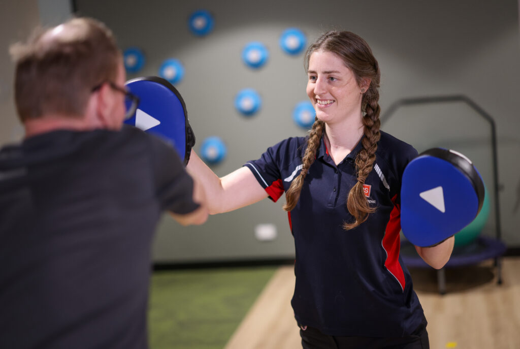 A neurological physio therapist with braided hair smiling while holding blue training pads for a patient to strike.