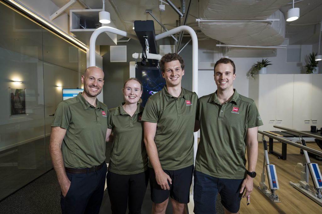 Four staff members in matching olive-green polo shirts with MS Queensland logos stand together in a physical therapy clinic.