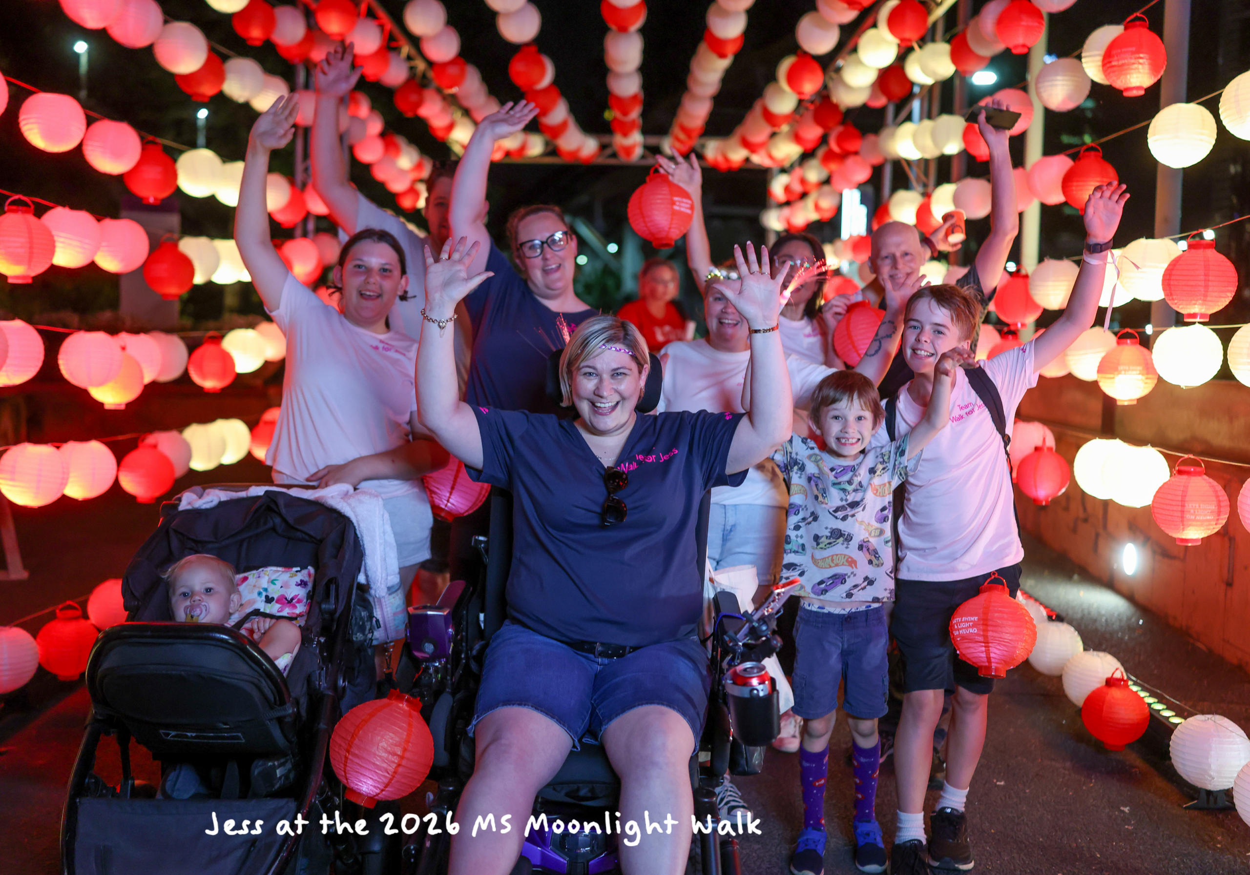 A diverse group, including a child in a stroller and a woman in a wheelchair, smiling and waving under a canopy of glowing red and white lanterns while living with MS at the 2026 Moonlight Walk.